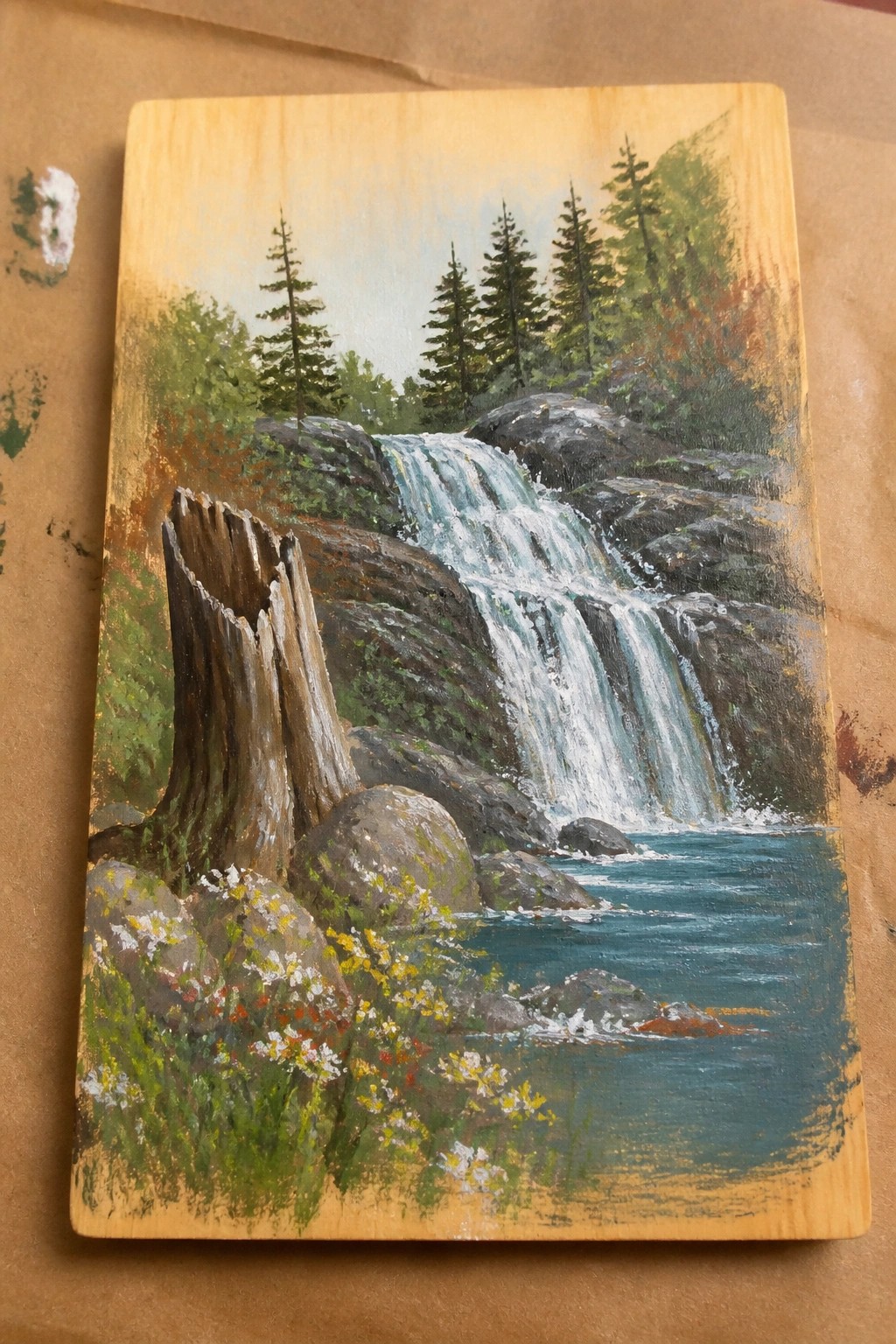 Wooden panel painting of cascading waterfall amid rocks, pines, and wildflowers