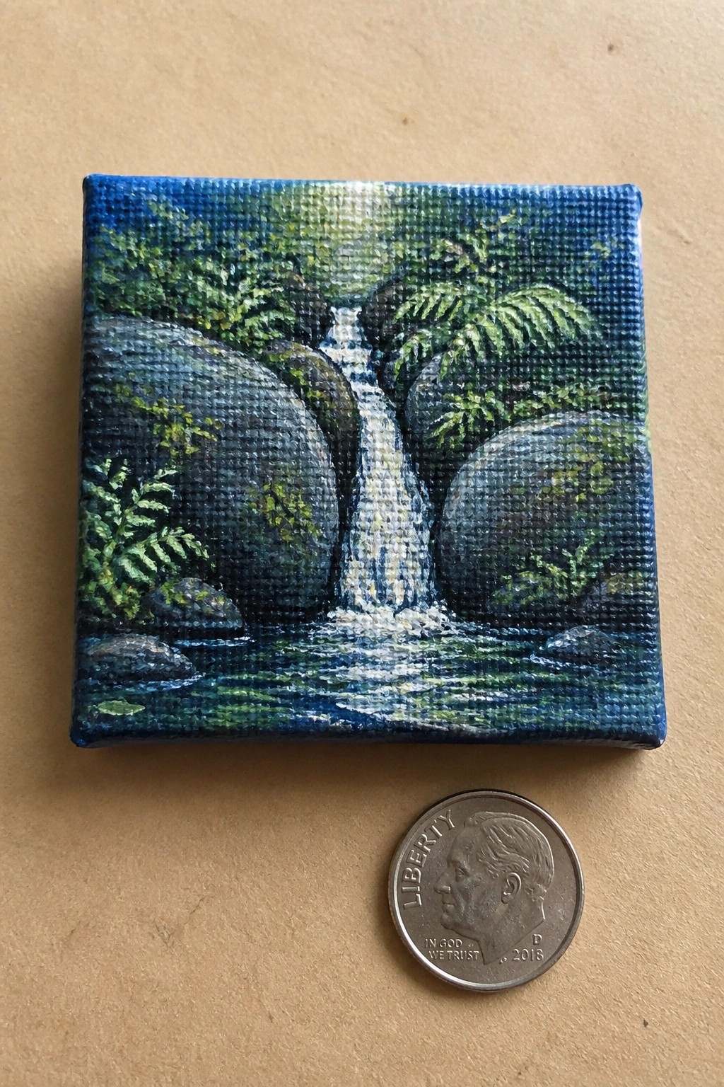 Tiny canvas painting of waterfall between rocks with ferns beside a dime