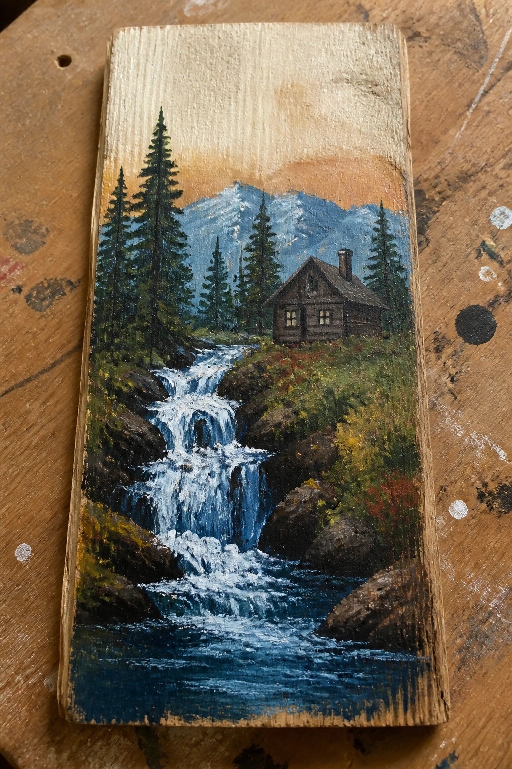 Landscape painting on wood of log cabin by rocky waterfall with mountains and pines
