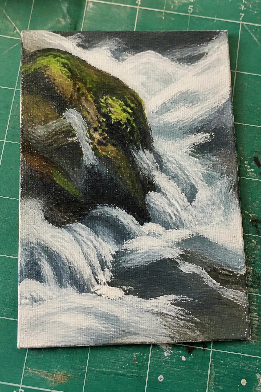 Acrylic painting of white water cascading over dark mossy rocks on a green cutting mat.