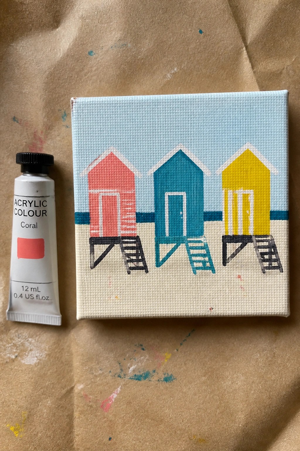Coral acrylic paint tube beside canvas with red, teal, and yellow beach huts