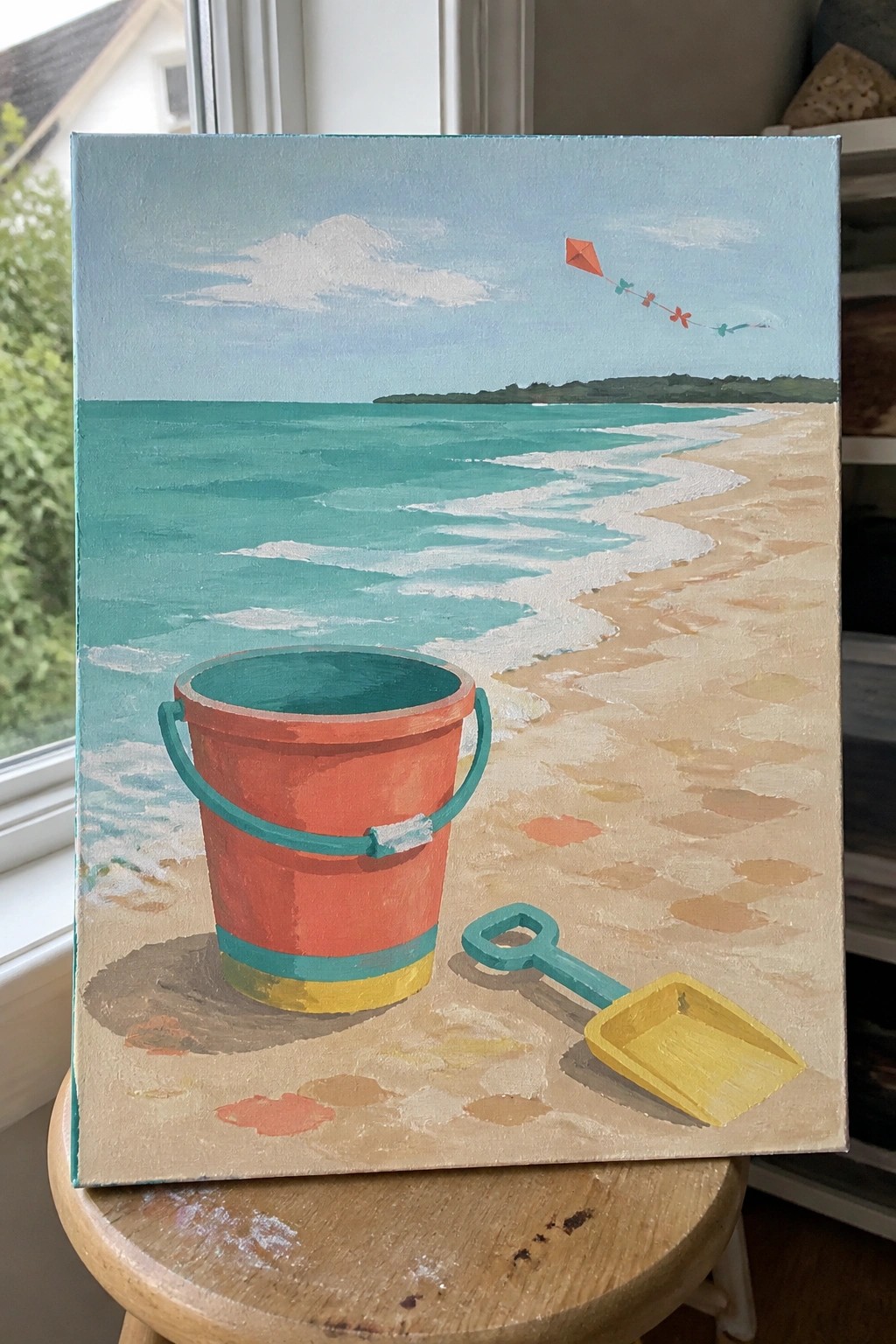 Red bucket and yellow shovel on sandy beach with turquoise waves and flying kite.