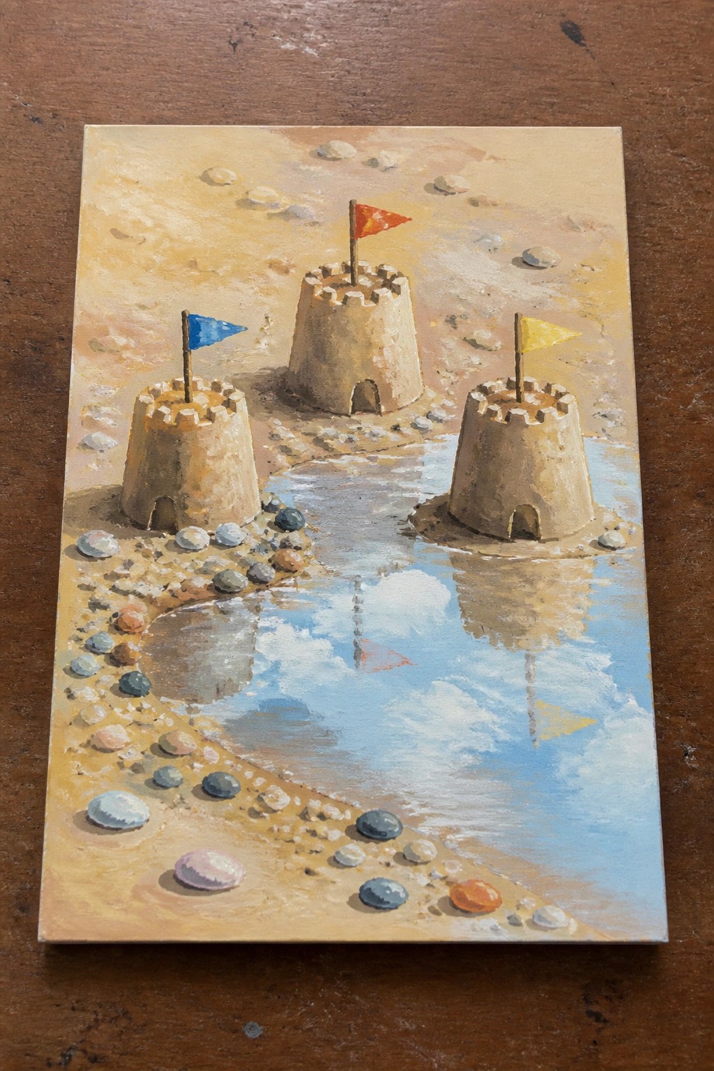 Three sandcastles with blue, red, and yellow flags beside a sky-reflecting puddle on sand.