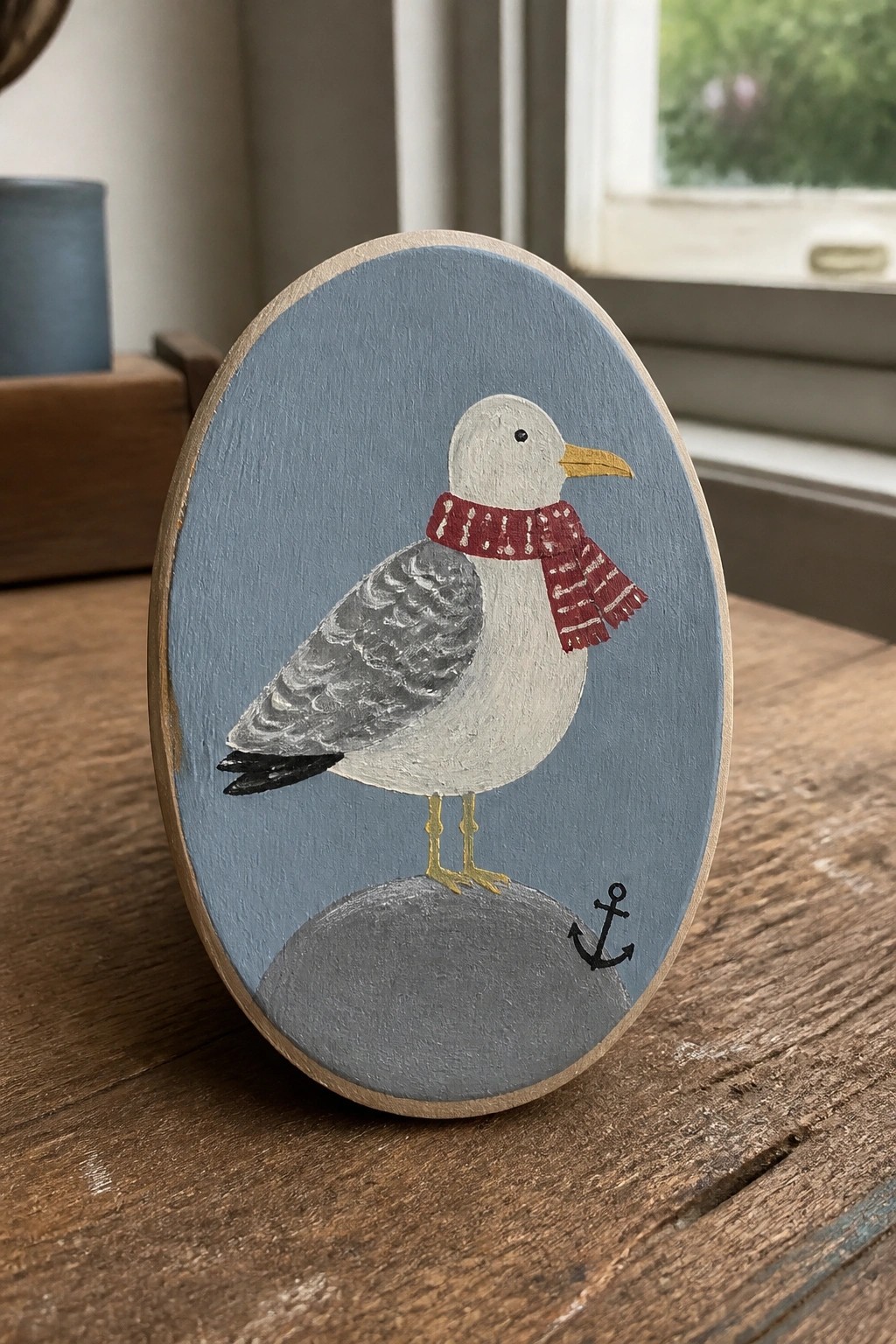 Wooden oval plaque with seagull in red scarf on rock beside black anchor