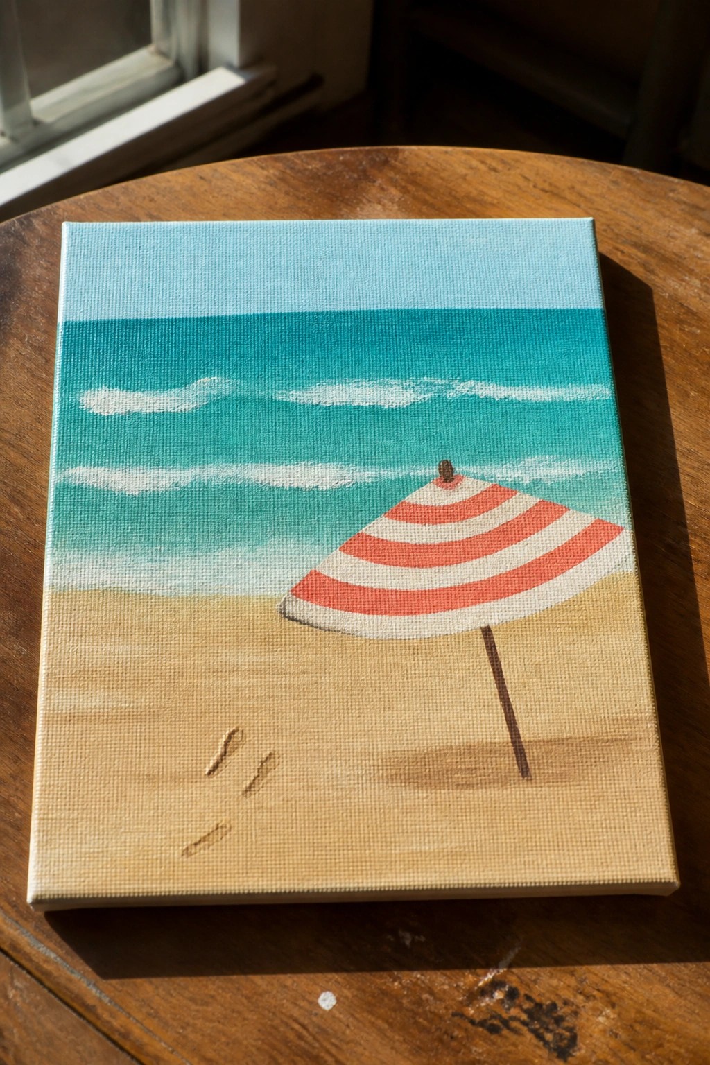 Beach painting with red-striped umbrella on sand, turquoise waves, and footprints
