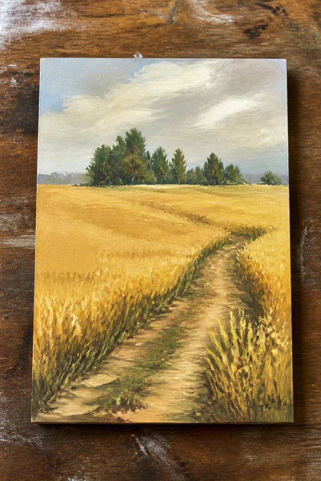 An acrylic painting of a dirt path winding through a golden wheat field toward a row of green trees under a cloudy blue sky, displayed on a wooden surface.