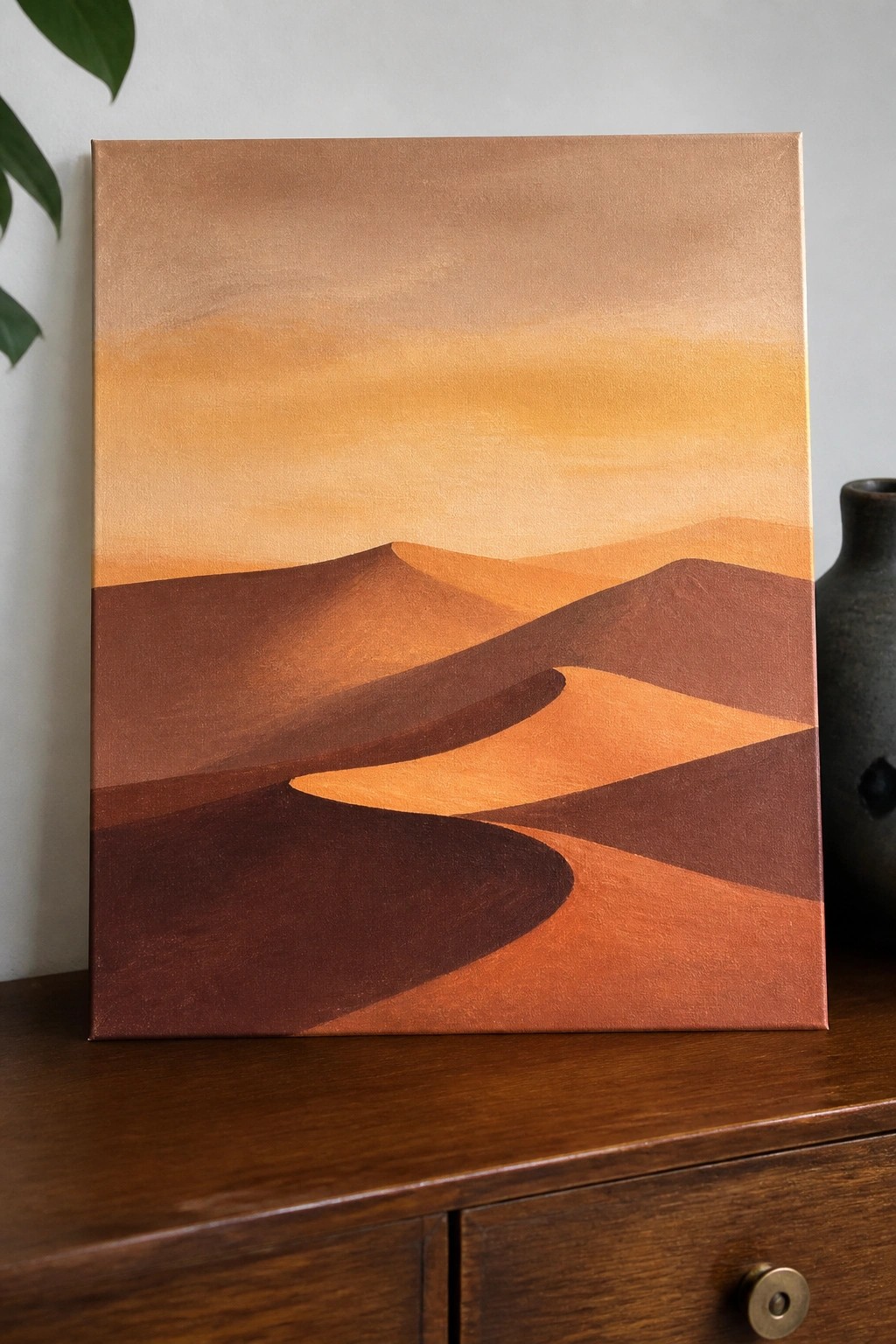 Acrylic painting of stylized sand dunes in shades of brown and orange with a gradient yellow-orange sky on a canvas resting on a wooden dresser.