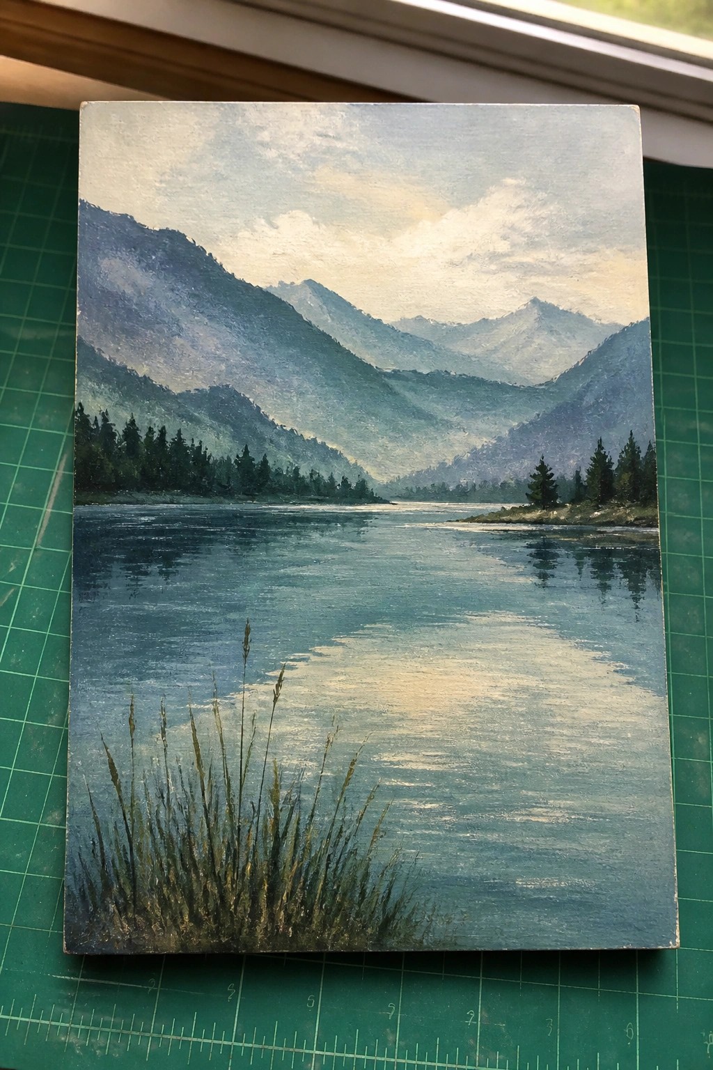 Painting of misty mountains reflected in calm lake with pine trees and foreground grass.