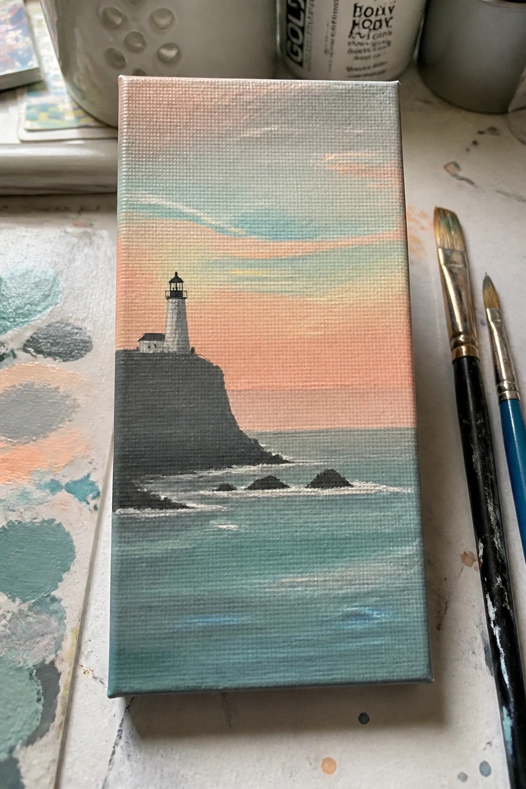 Small canvas shows lighthouse on cliff over ocean with pink sky, brushes and palette nearby.