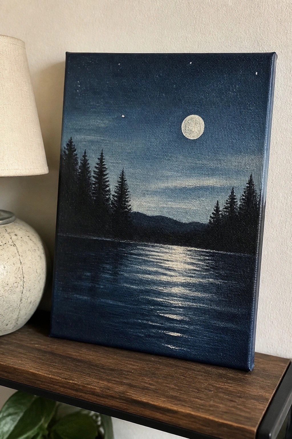 A rectangular acrylic painting of a dark blue night sky with a full moon, stars, silhouetted pine trees on a hill, and their reflection in a lake below, placed on a wooden shelf next to a lamp.