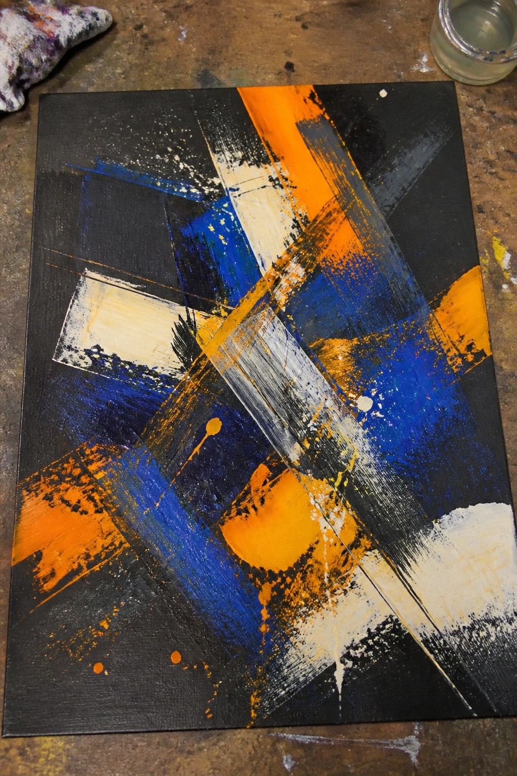 Abstract acrylic painting featuring thick orange, blue, and white brush strokes crossing a black canvas with visible splatters and overlaps.
