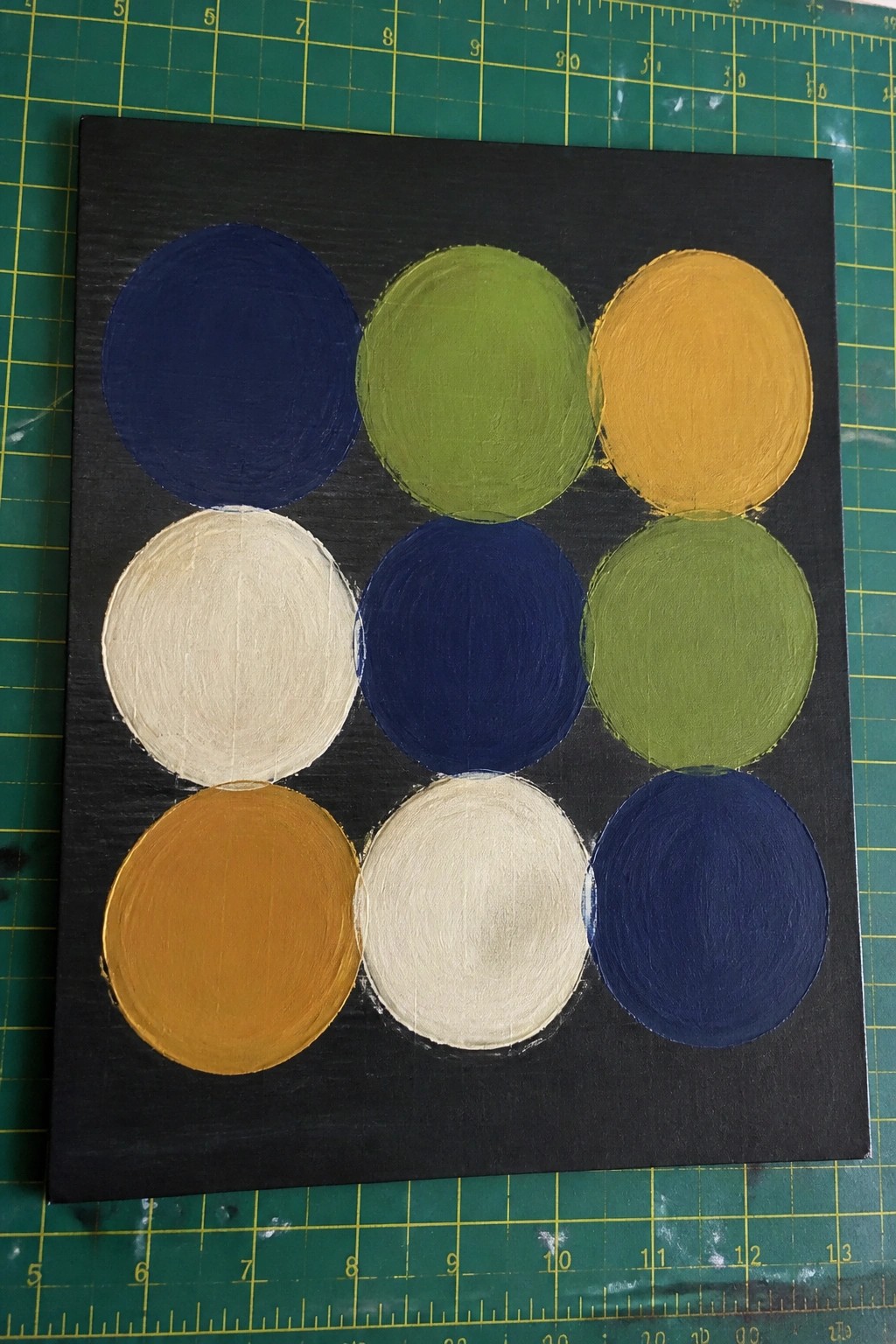 Nine colorful circles painted in a grid on black square canvas over green mat