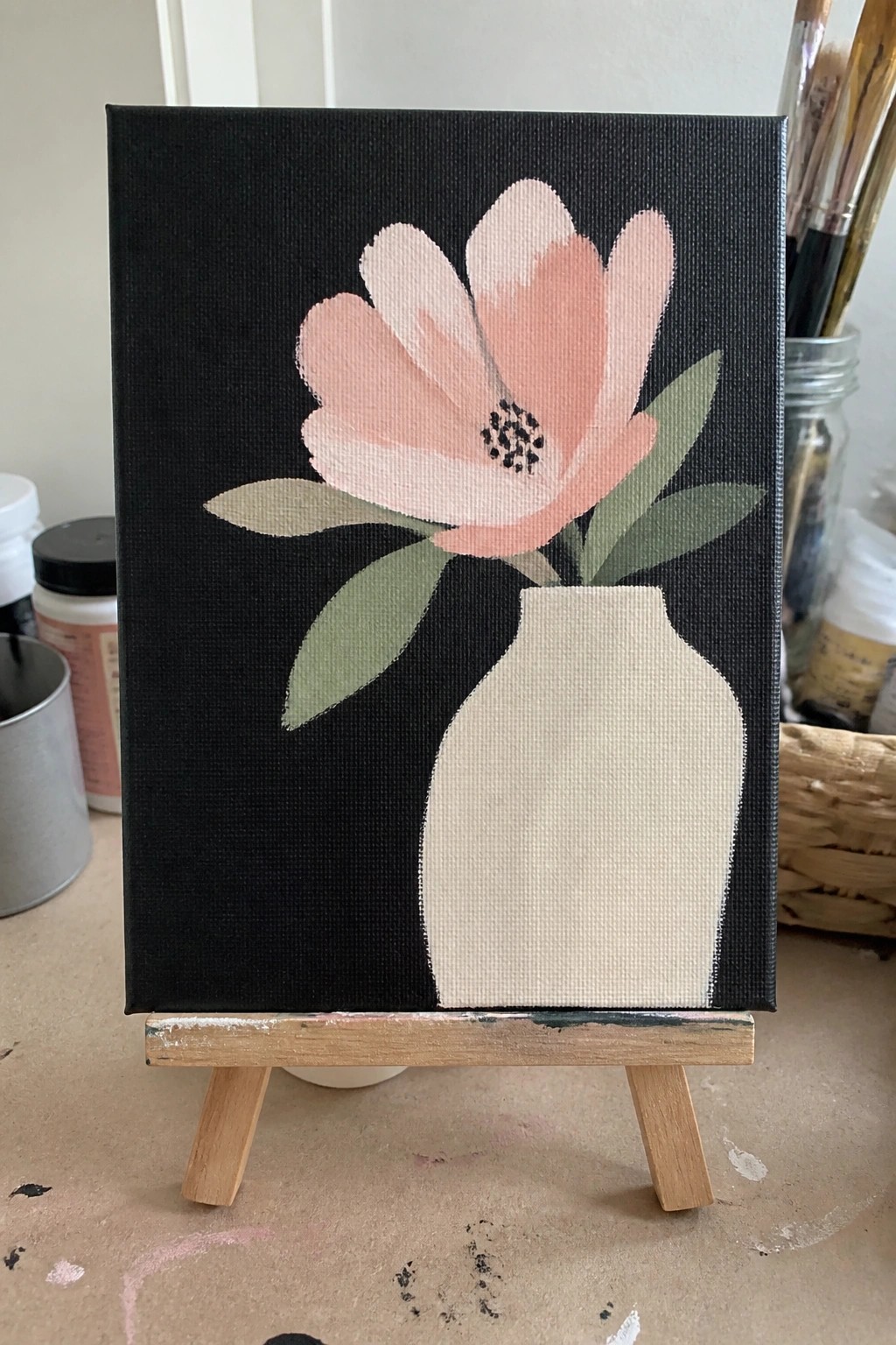 Pink flower with green leaves in cream vase on black canvas atop wooden easel