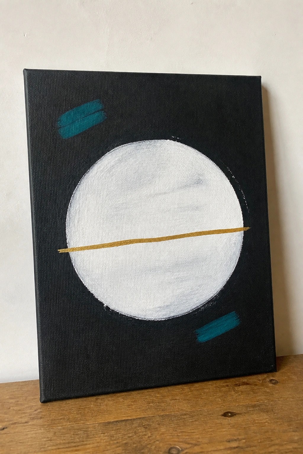 Abstract painting: white circle with gold line on black canvas, teal accents on wood.