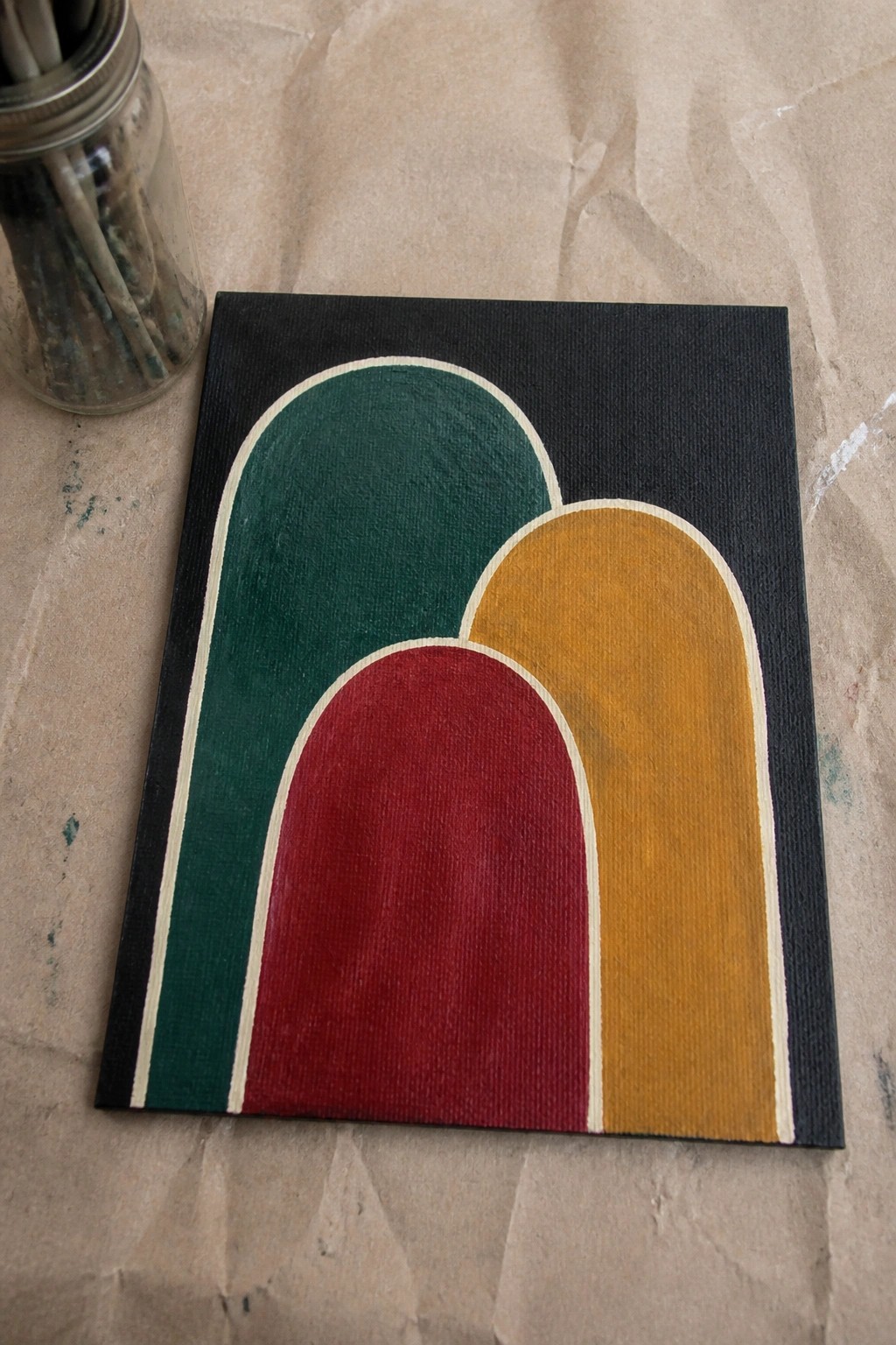 Abstract painting with overlapping green, red, and yellow arches on black canvas beside paintbrush jar.