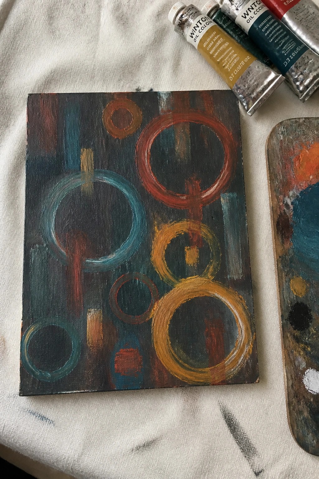 An abstract oil painting with overlapping circles and rings in teal, red, orange, and yellow against a dark background, shown beside oil paint tubes and a palette.