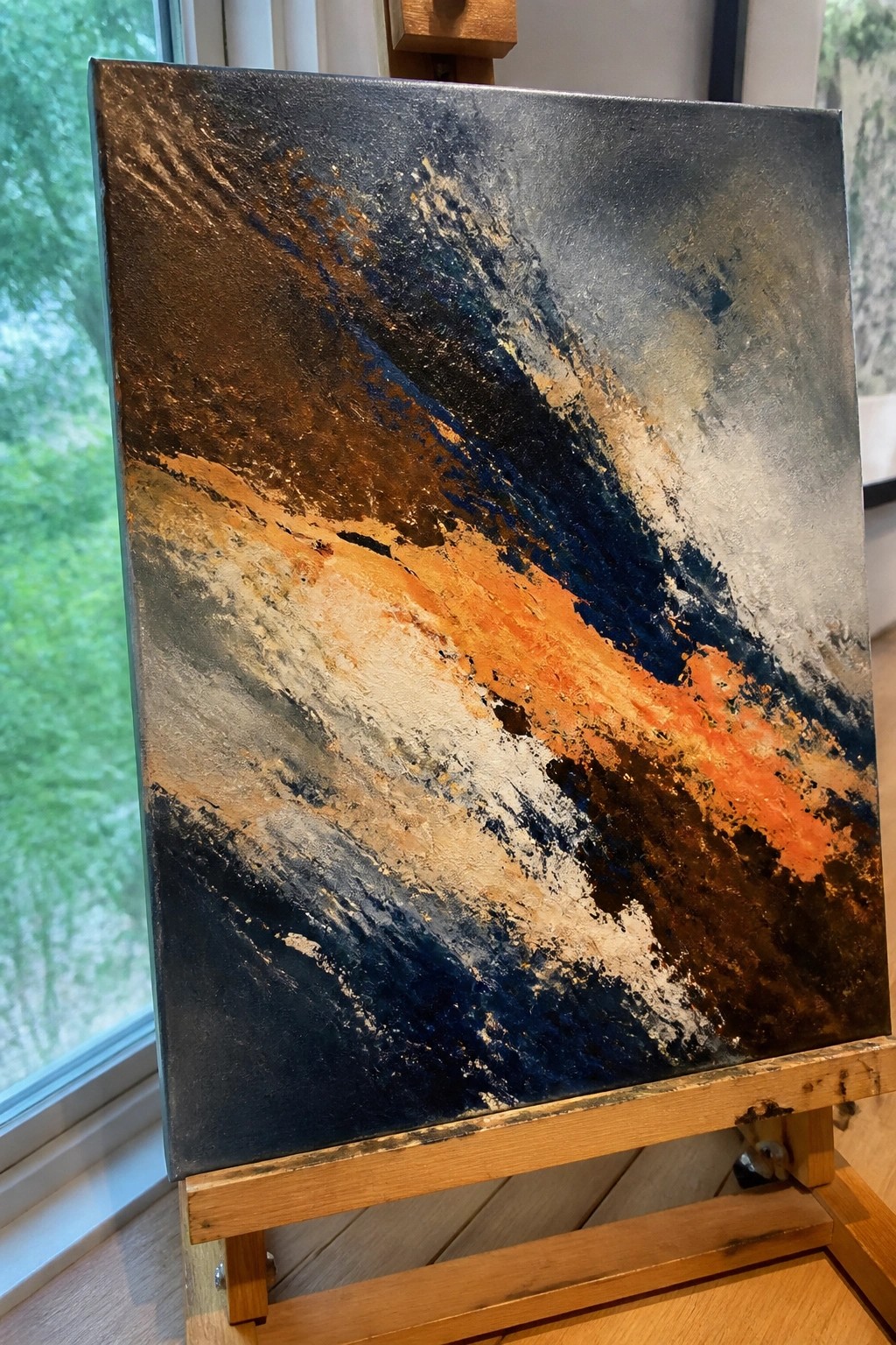 Abstract painting with bold orange, blue, and black brushstrokes on wooden easel near window