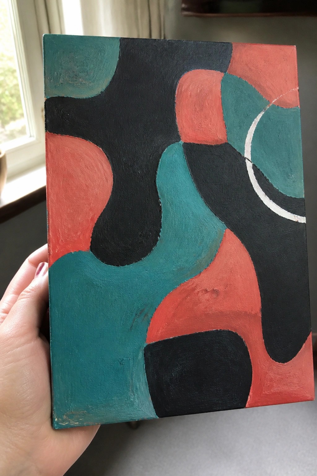 Hand holding abstract painting with teal, black, coral shapes and white arc.