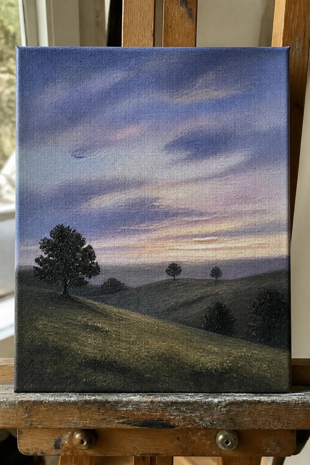 Landscape painting on easel with rolling hills, silhouetted trees, and colorful dusk sky.
