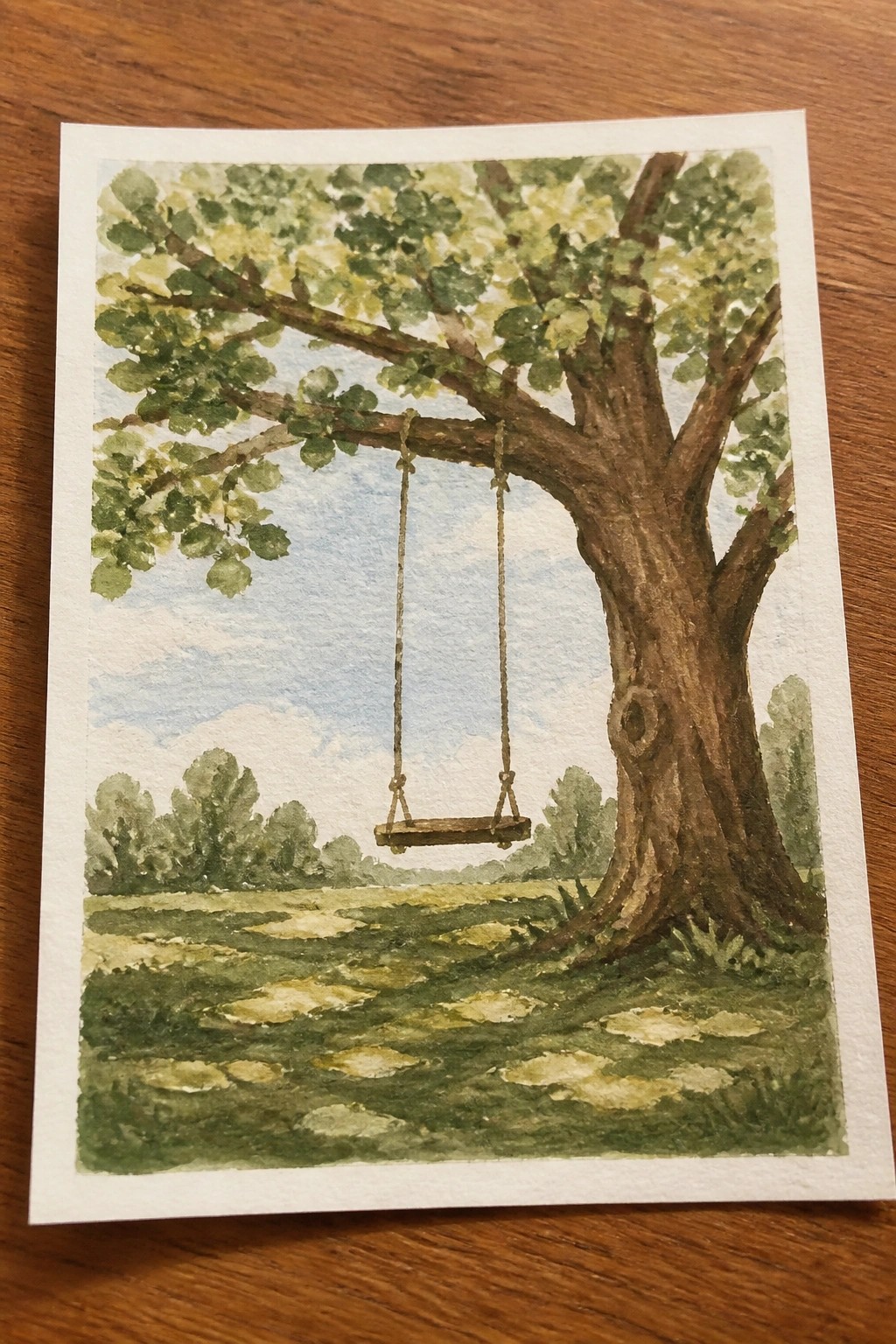 Watercolor painting of a rope swing hanging from a leafy tree in a green field.