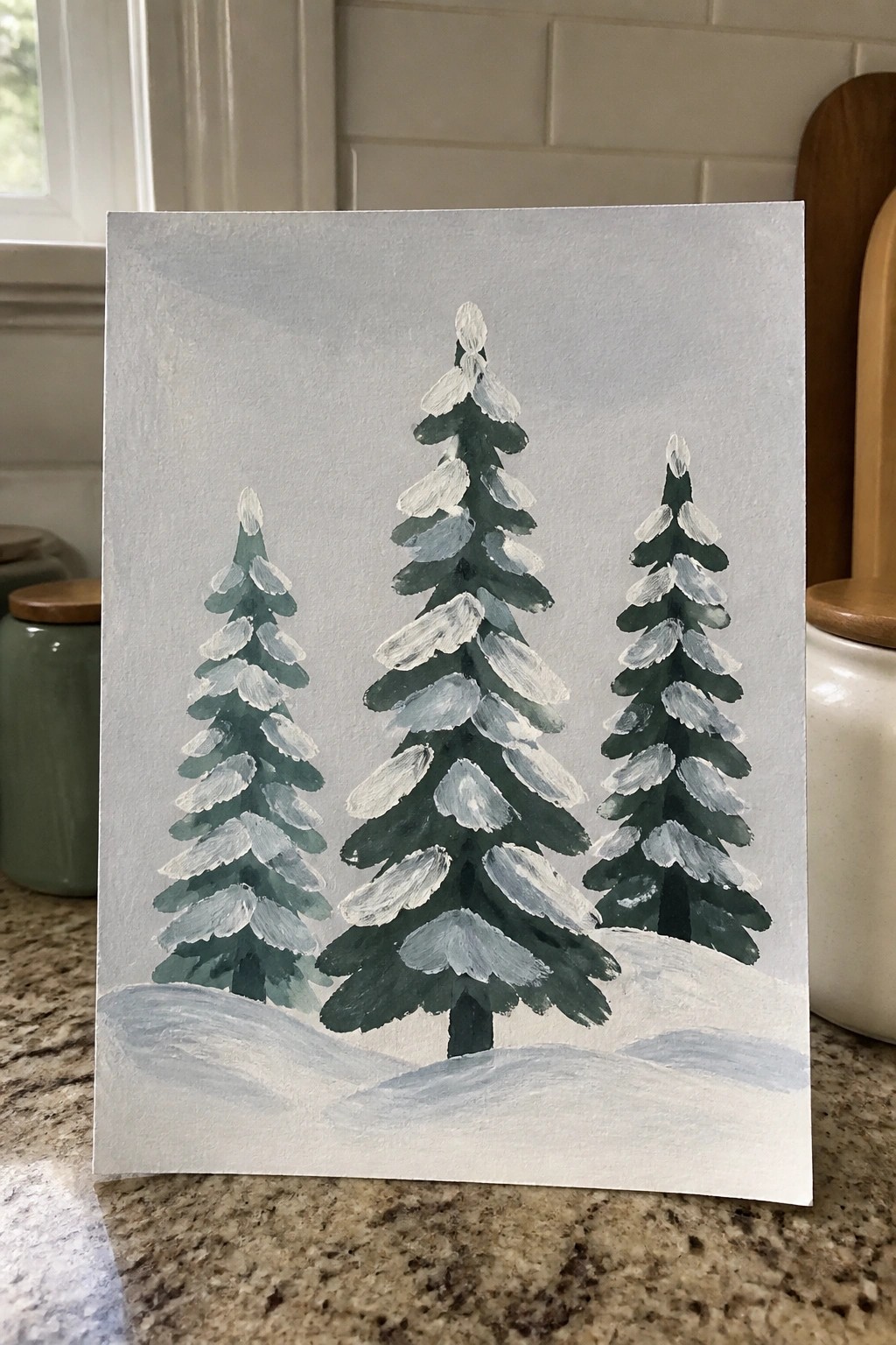 Three snow-covered pine trees painted in watercolor on light gray paper.