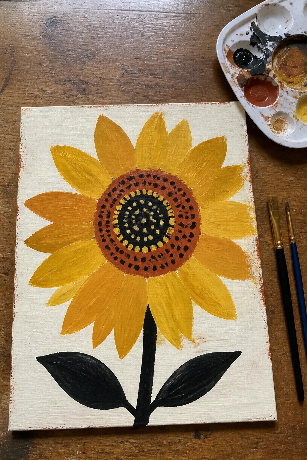 Yellow sunflower painting on canvas beside paint palette and brushes on wood table