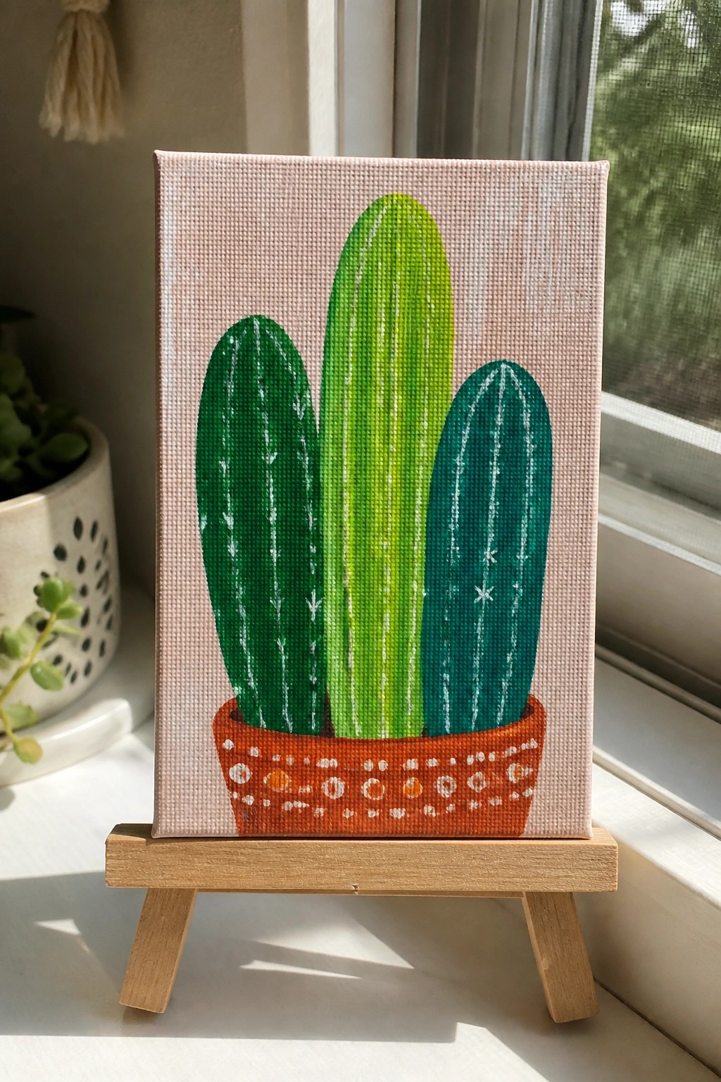 Three green cacti in patterned terracotta pot on pink canvas with wooden easel.