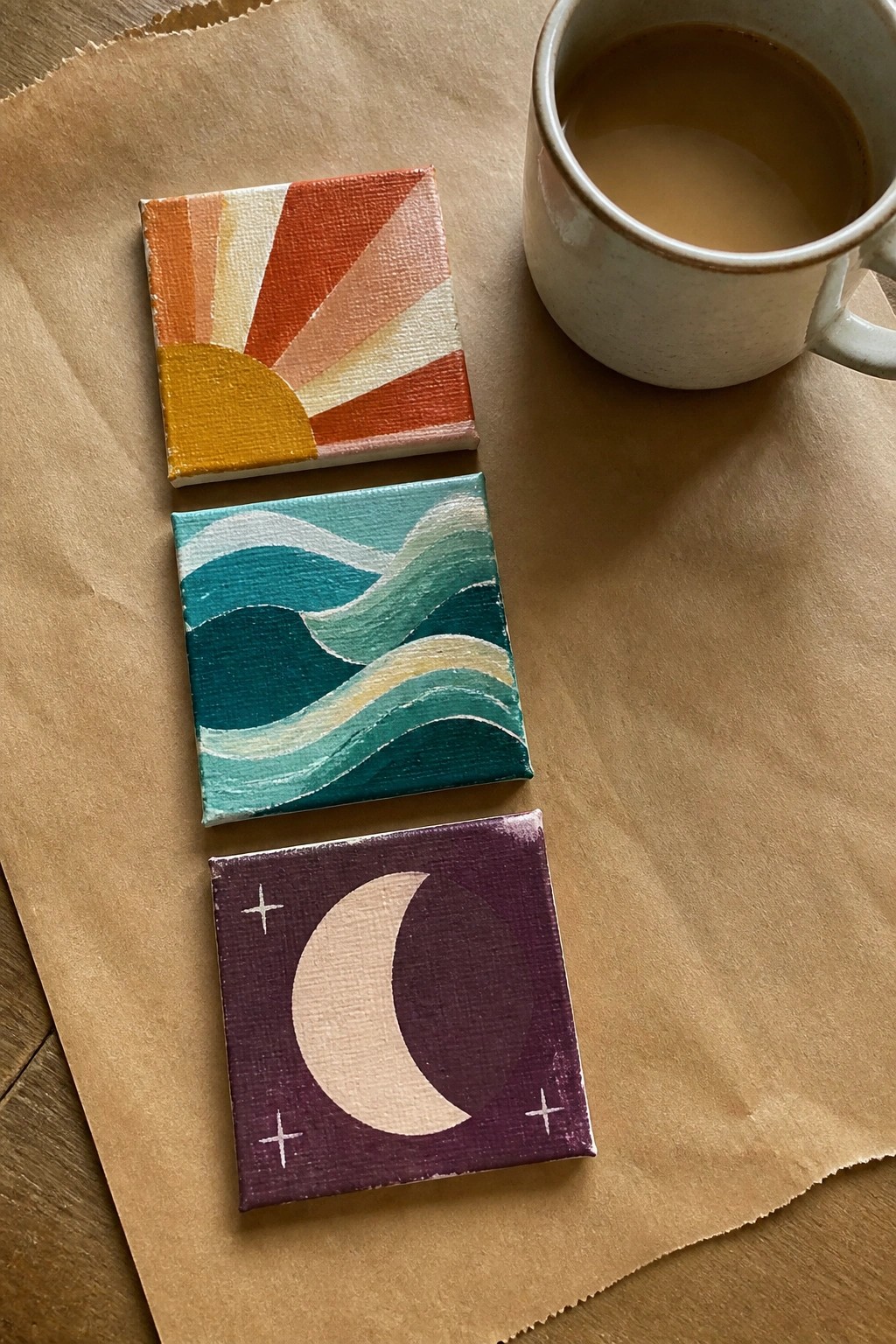 Three mini canvases with sunset, waves, and moon paintings beside a coffee mug on paper