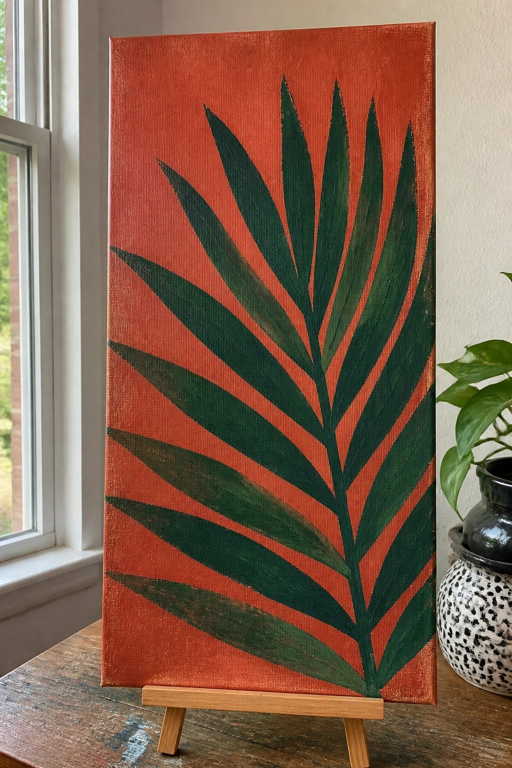 Orange canvas with large green palm leaf painting on wooden easel beside plant.