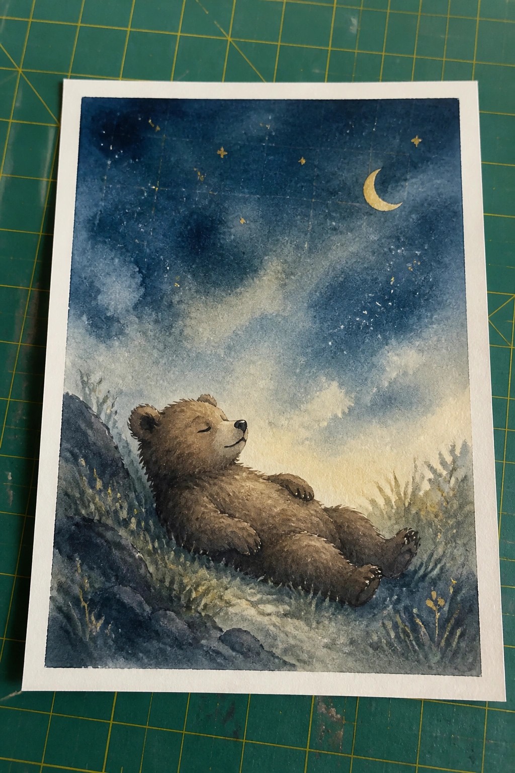 Watercolor painting of a sleeping brown bear resting under starry night sky with crescent moon.