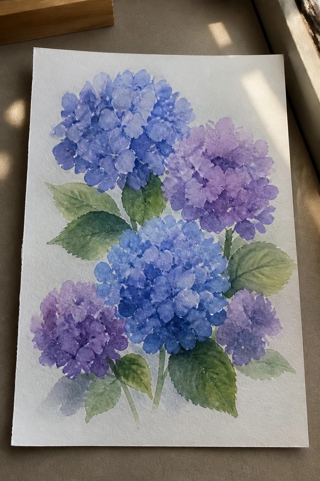 Watercolor painting of blue and purple hydrangea flower clusters with green leaves