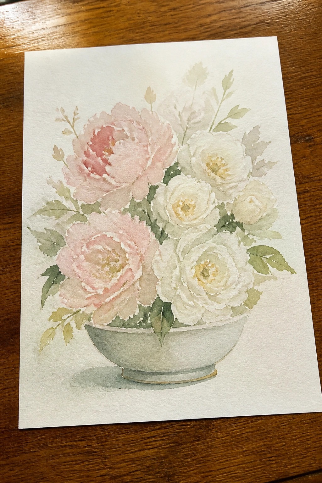 Watercolor of pink and white peonies with green leaves in a light bowl on wood