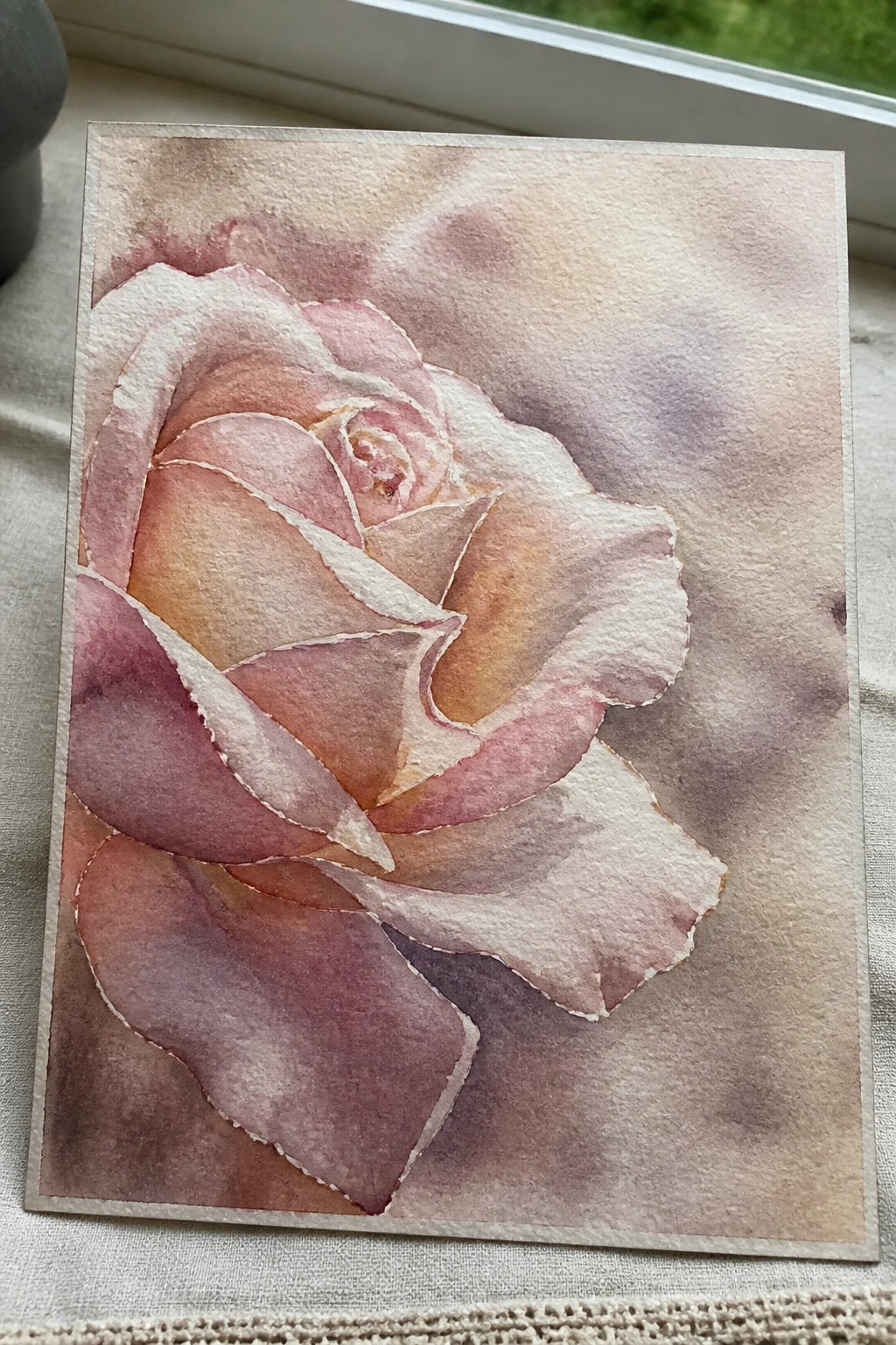 Watercolor painting of a blooming rose in soft pink, peach, and purple tones on a windowsill.