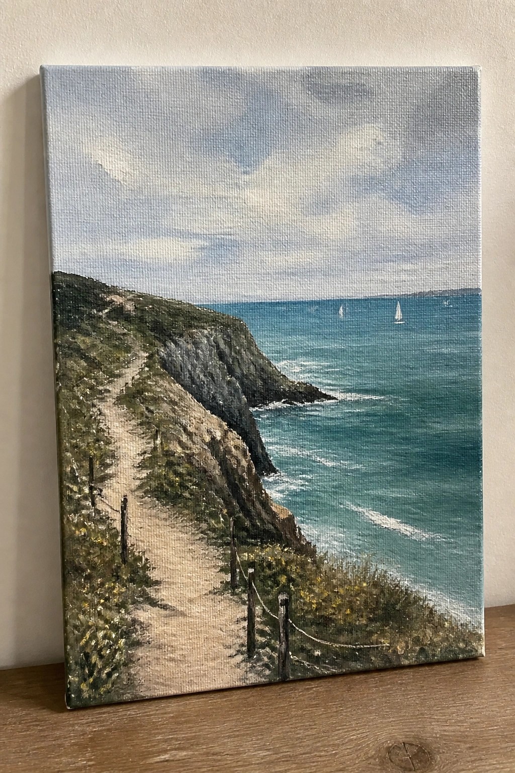 Oil painting of cliffside path above teal ocean with sailboats under cloudy sky.