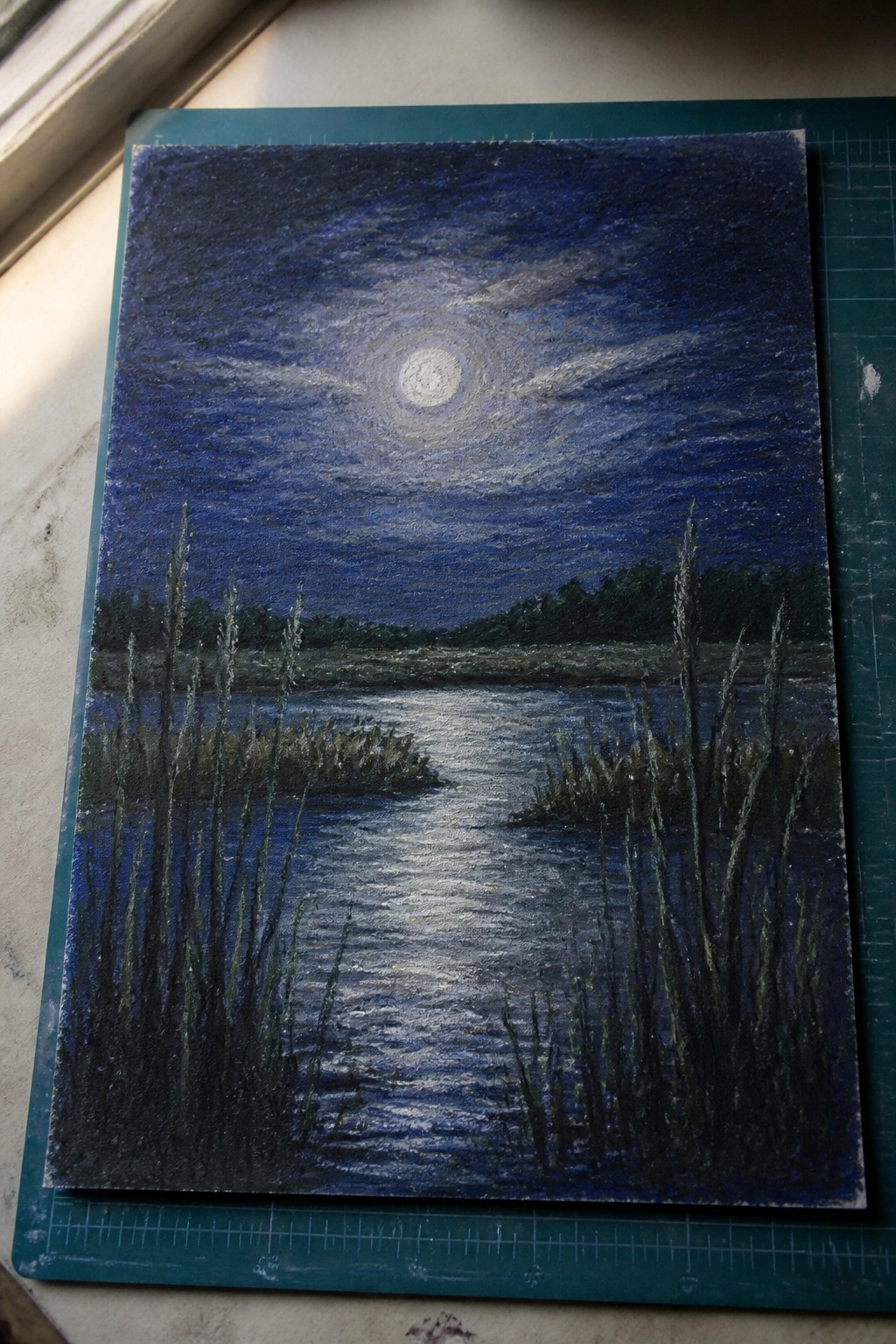 A painting of a full moon reflecting on a dark river at night, with tall reeds in the foreground and trees along the distant shore, resting on a green cutting mat.