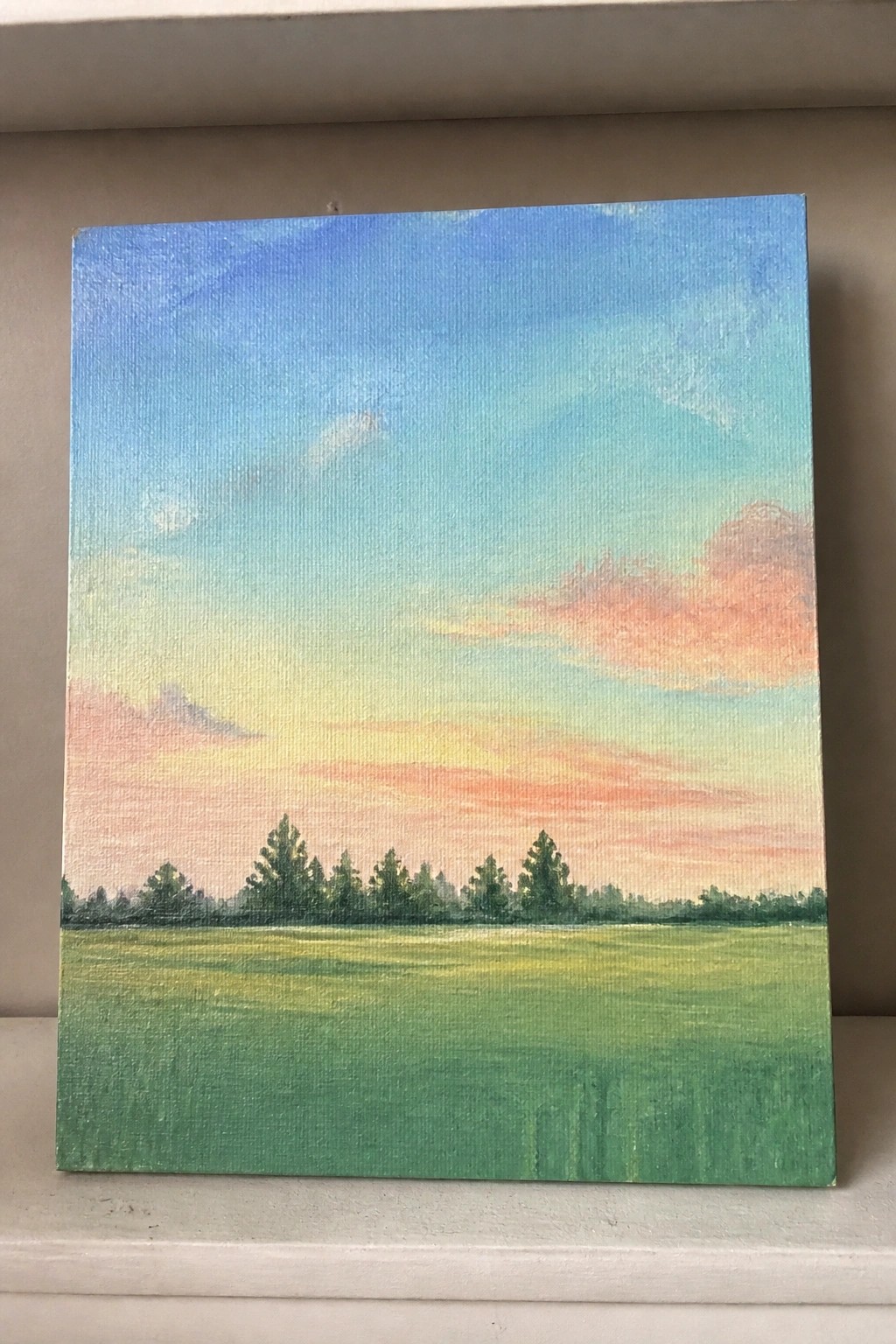 Landscape painting on canvas shows green field, trees, and vibrant sunset sky.