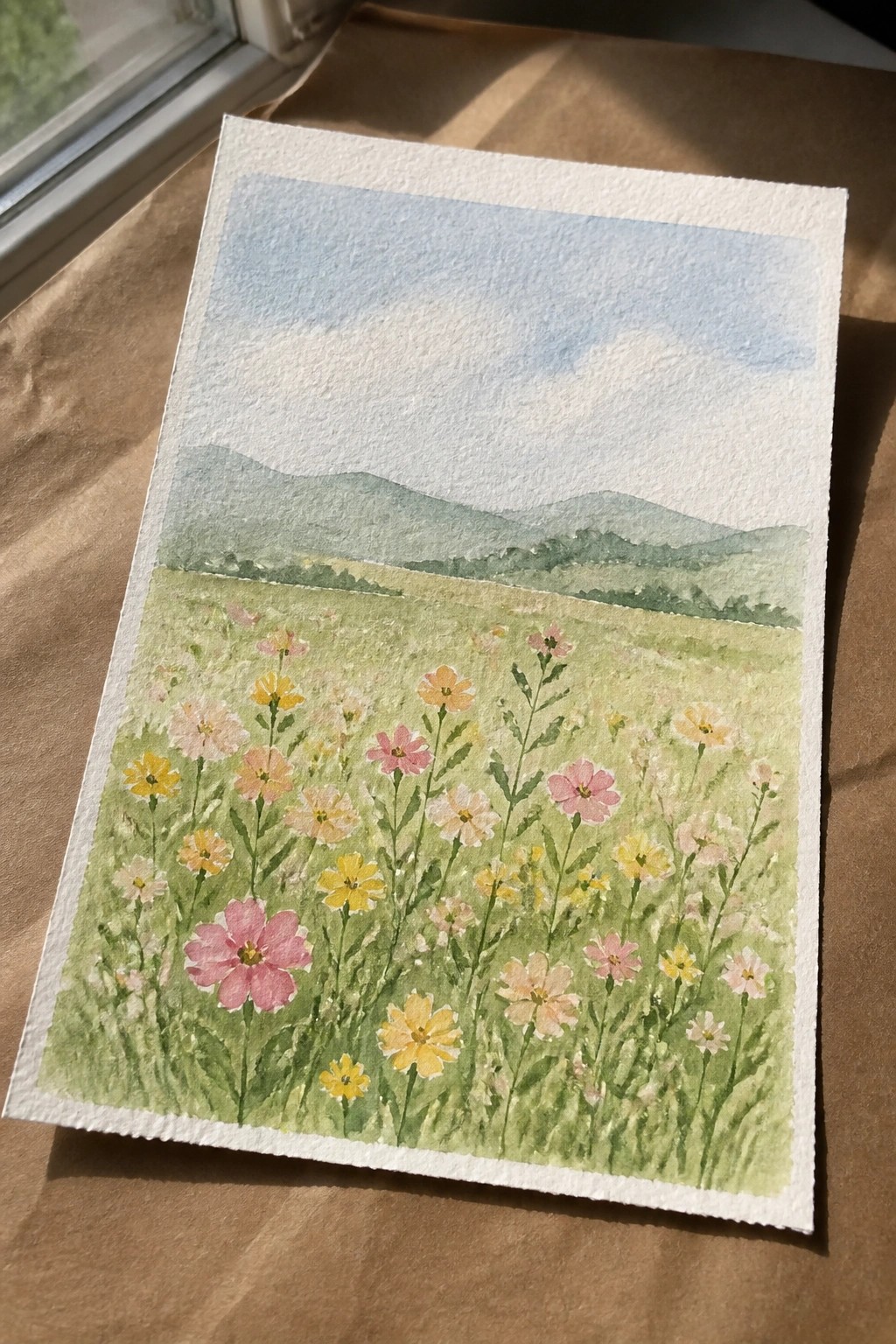 A watercolor painting of a green meadow filled with pink, yellow, and white wildflowers, with layered hills and a blue sky in the background.