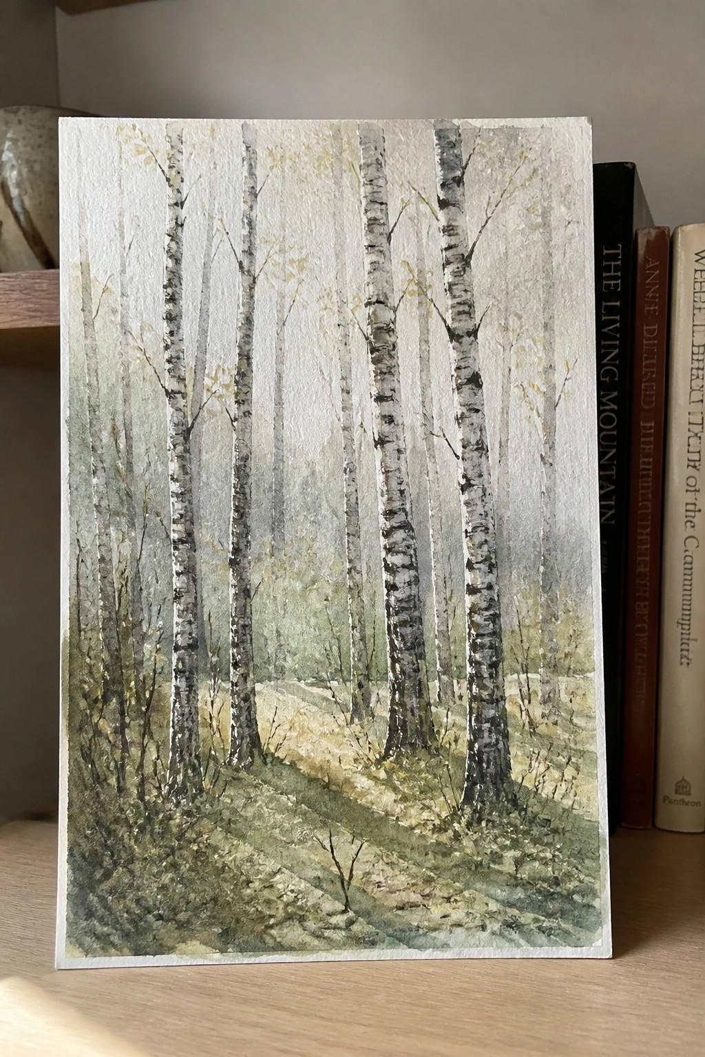 Watercolor painting of birch trees in misty forest beside books on wood shelf