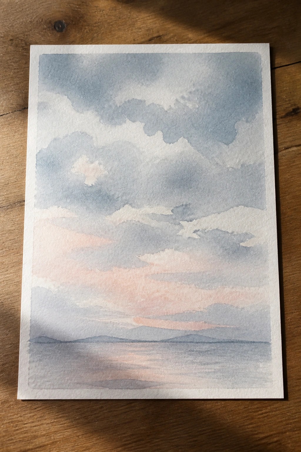 Watercolor painting of pink-blue cloudy sky over calm lake with distant hills on wood.