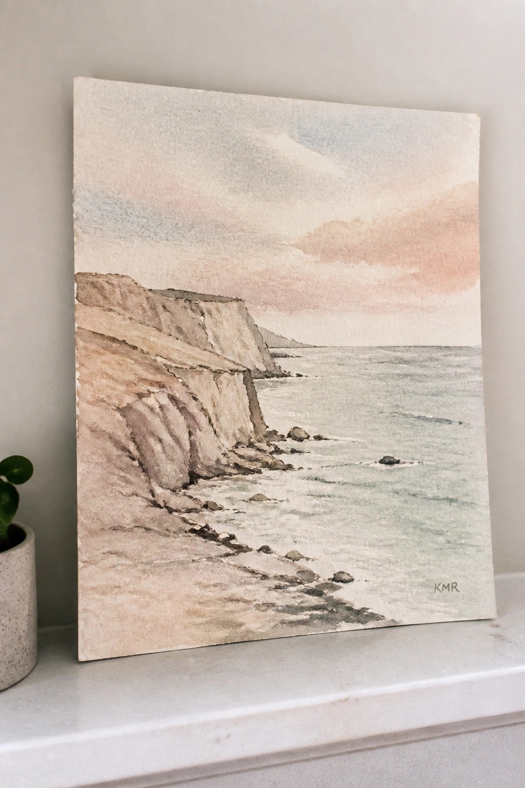 Watercolor of layered cliffs by the sea under a soft pastel sky beside a potted plant