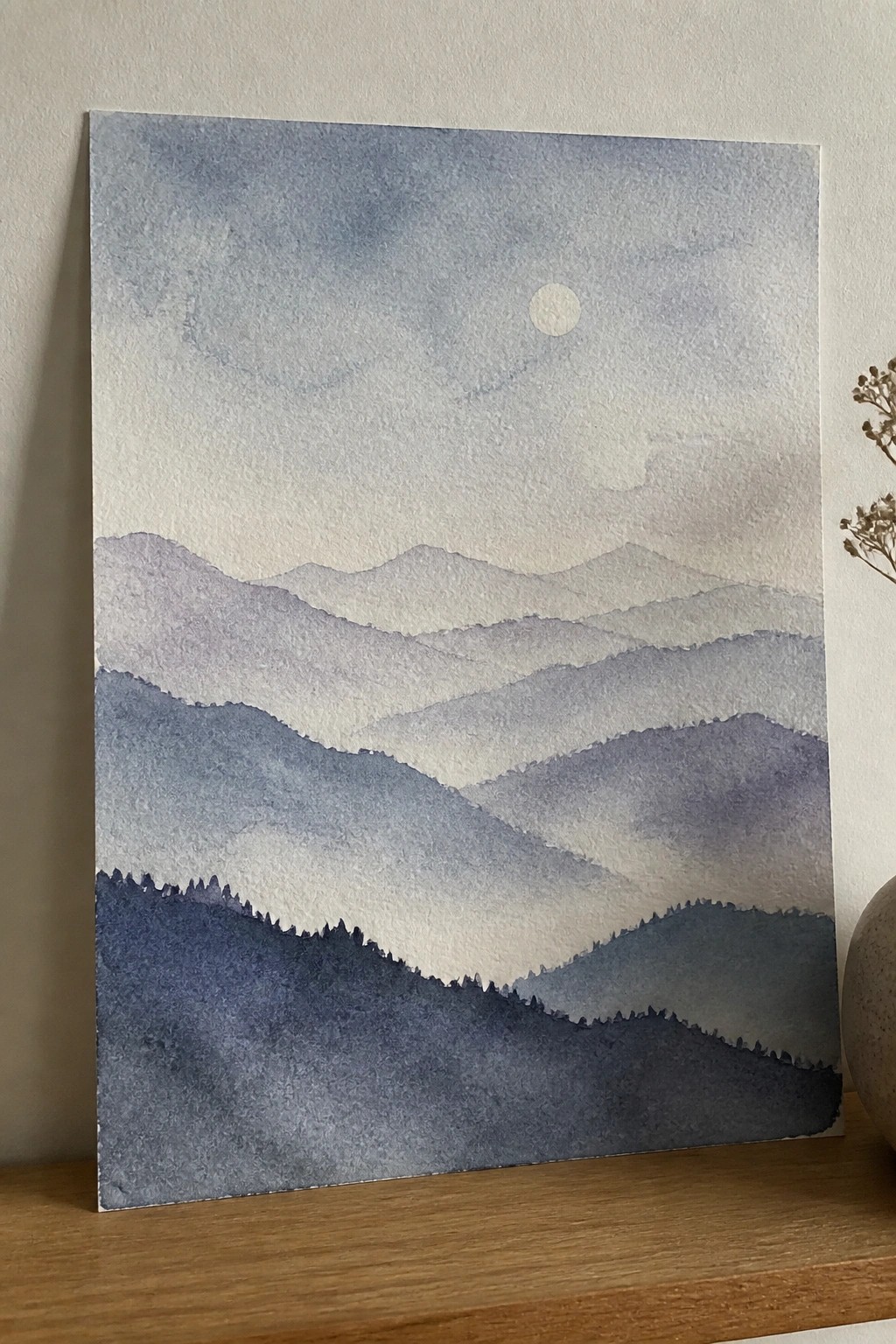 Layered blue watercolor mountains beneath pale moon on wooden shelf with vase
