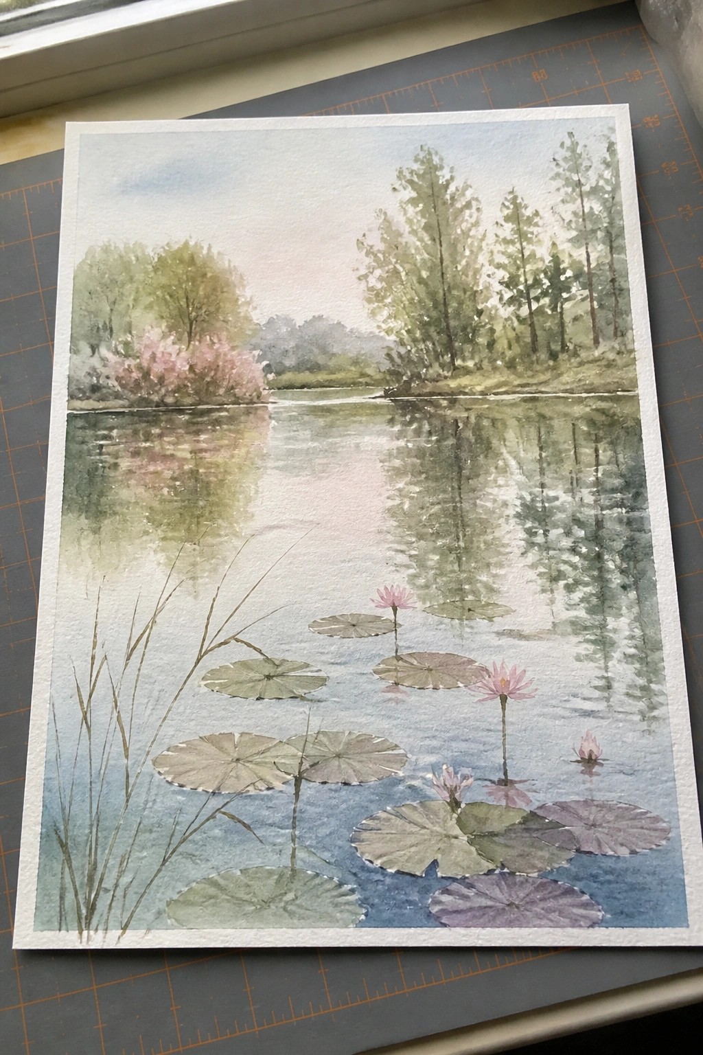 Watercolor landscape of a calm pond with pink lotus flowers, lily pads, and tree reflections.
