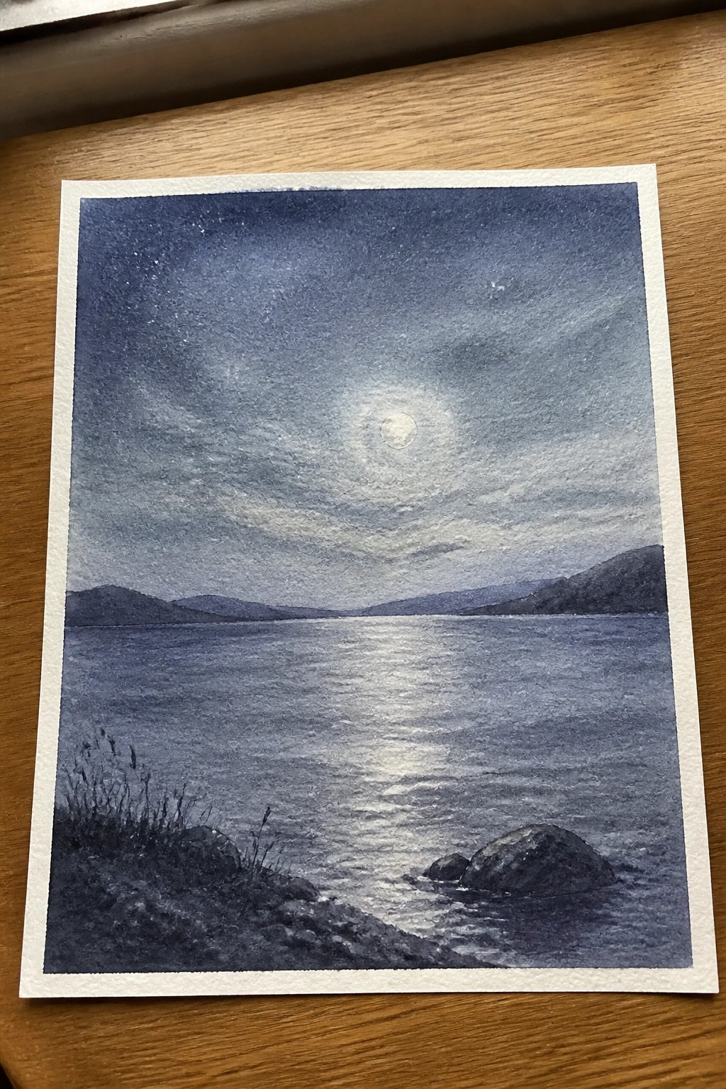 Watercolor night scene of full moon reflecting on calm lake with mountains and rocky shore
