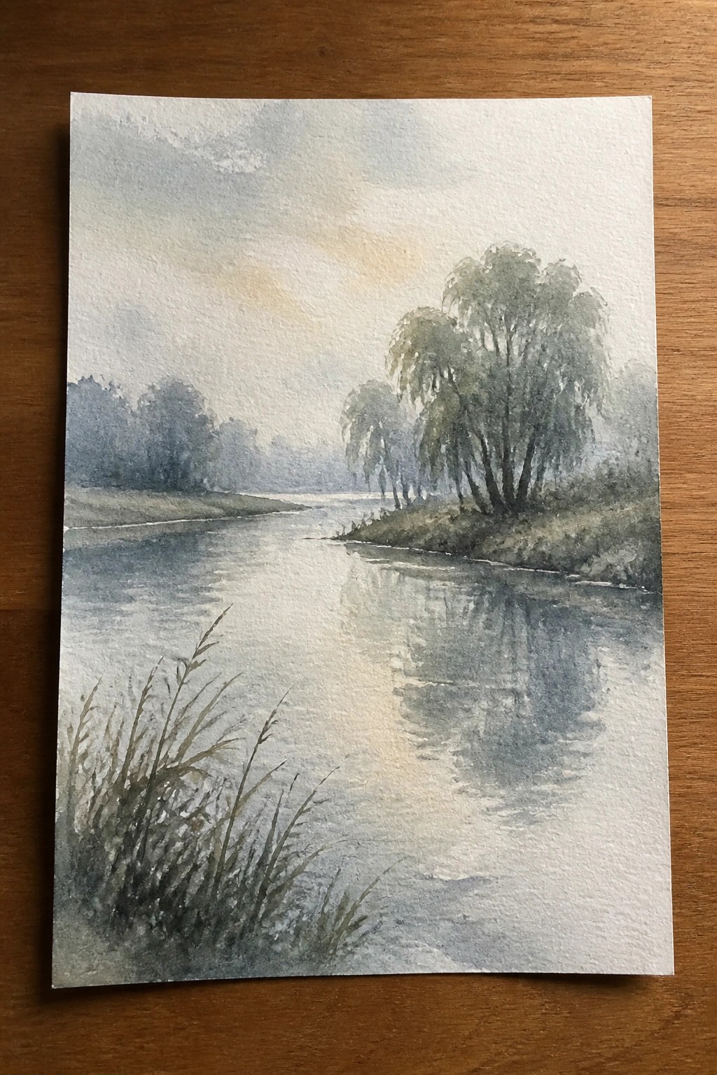 Watercolor of misty river with weeping willows reflecting on calm water and grass.