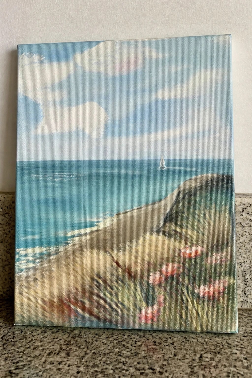 Canvas painting of blue ocean, sailboat, and pink flowers on grassy seaside cliff.