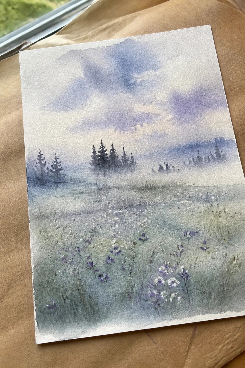 Watercolor painting of misty meadow with wildflowers, pine trees, and cloudy blue sky