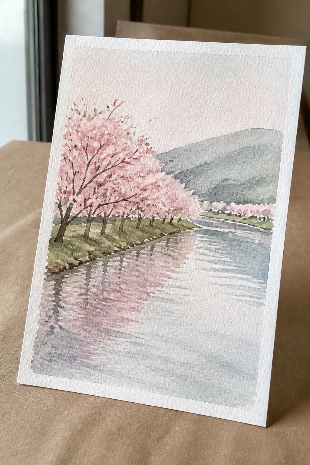 Watercolor of pink cherry blossoms along a reflective river with green mountains.