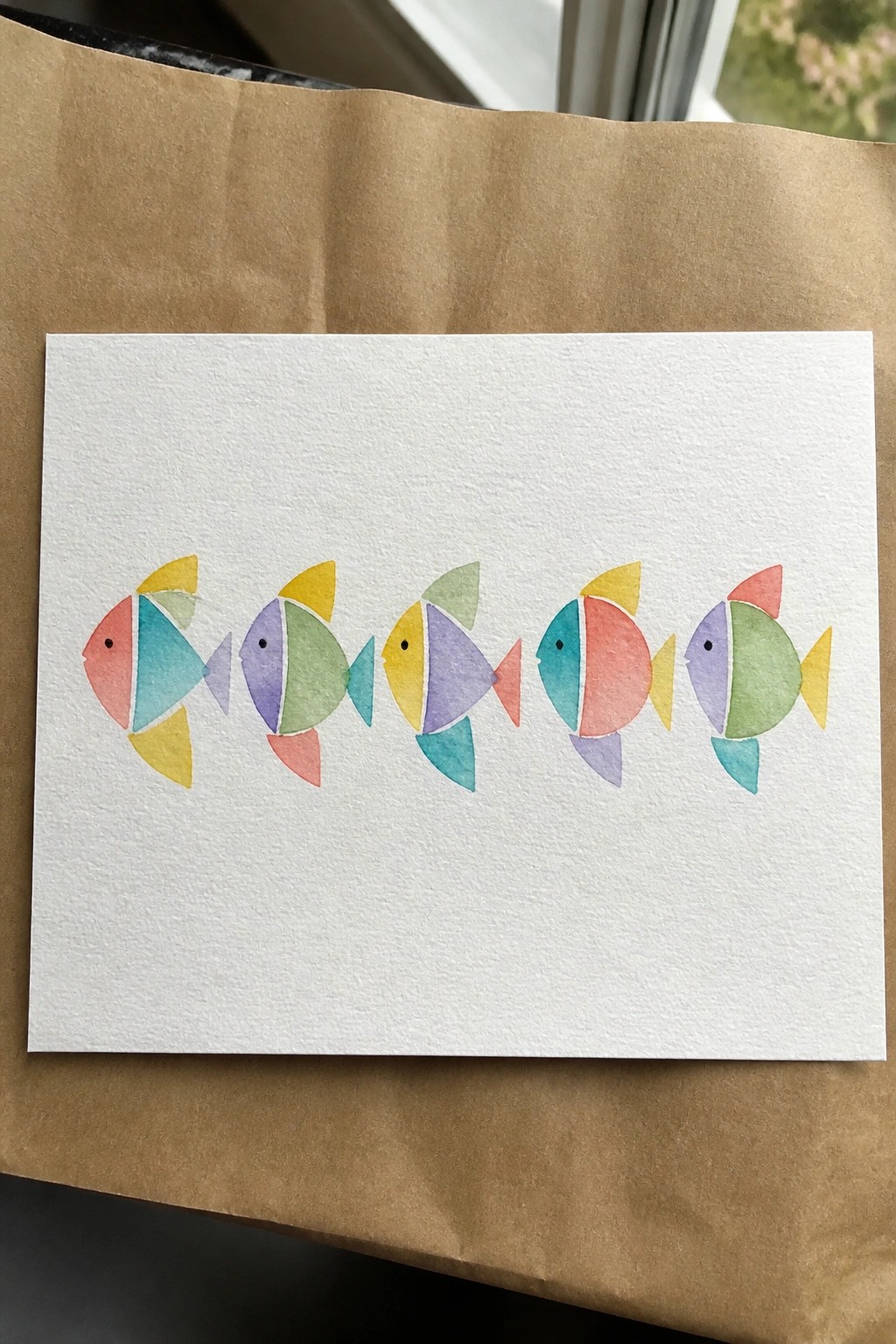 Five colorful geometric fish painted in watercolor on white textured paper.