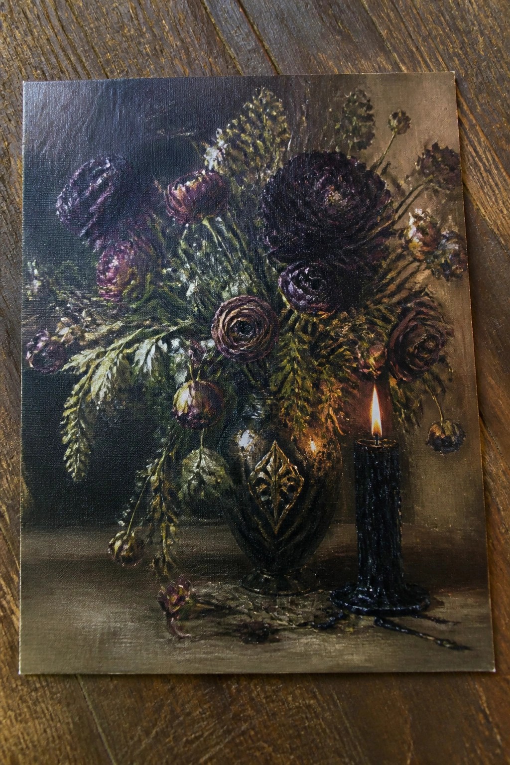 Dark purple flowers in ornate vase beside lit black candle on wood.