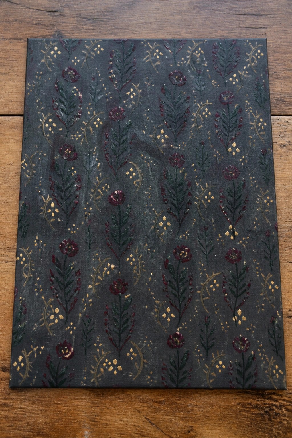 Dark canvas with maroon flowers, green stems, and gold accents on wood surface.